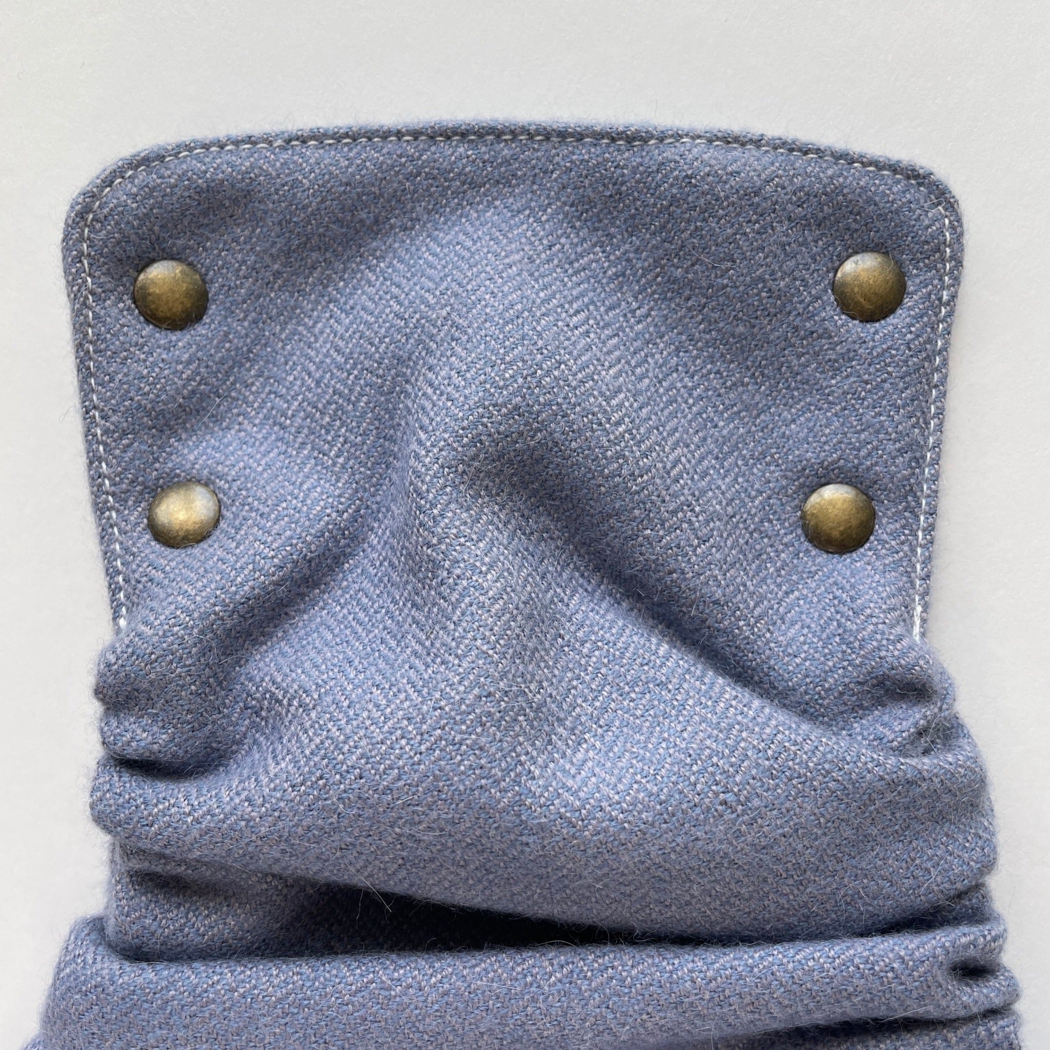 The front panel of a dawn-colored alpaca diaper cover with a blue-purple mini-herringbone pattern with 4 brass snaps in an antique finish laying on top of a white background.