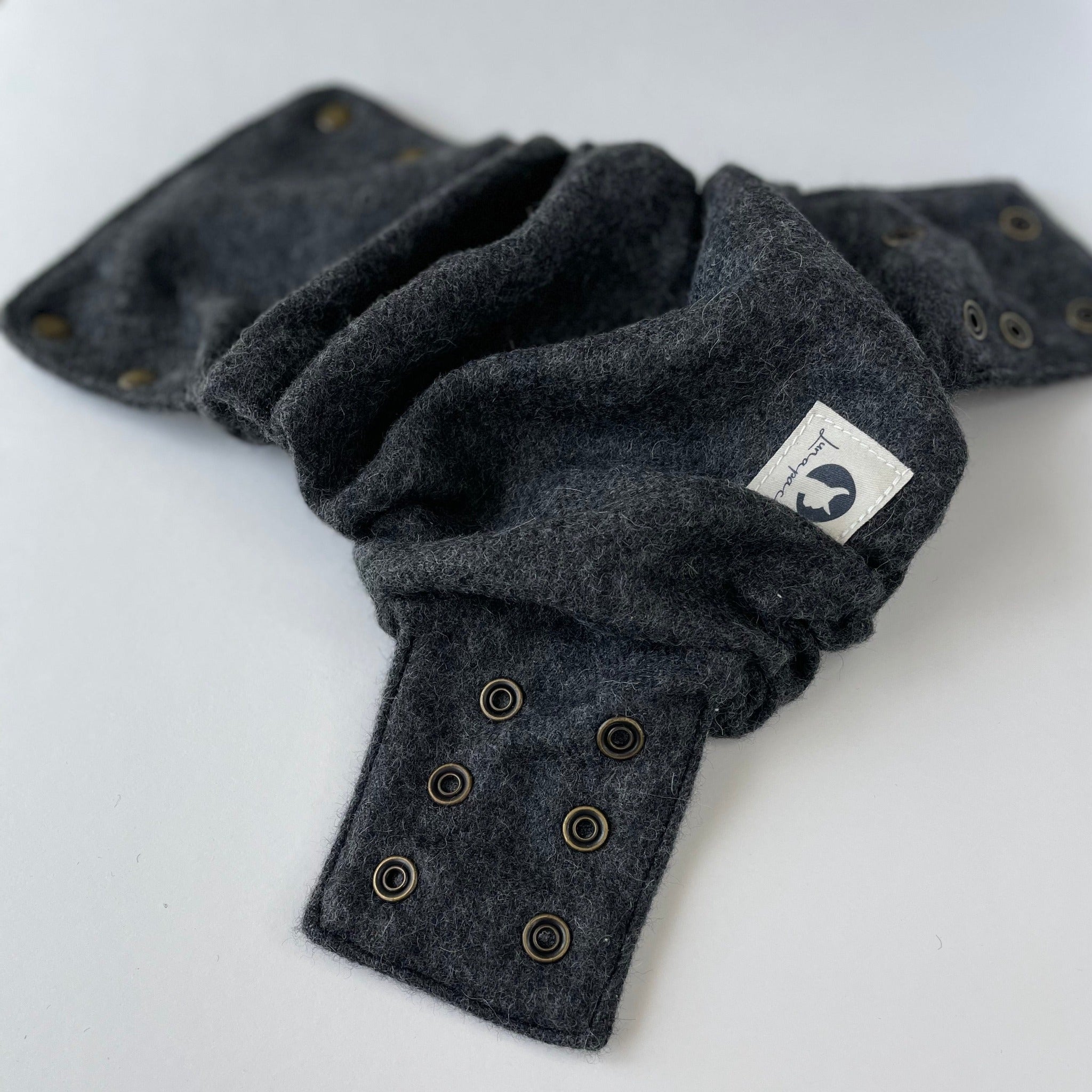A charcoal gray alpaca diaper cover laying open on a white background with 6 brass snap rings on the side panel and a square cotton logo tag in the foreground.