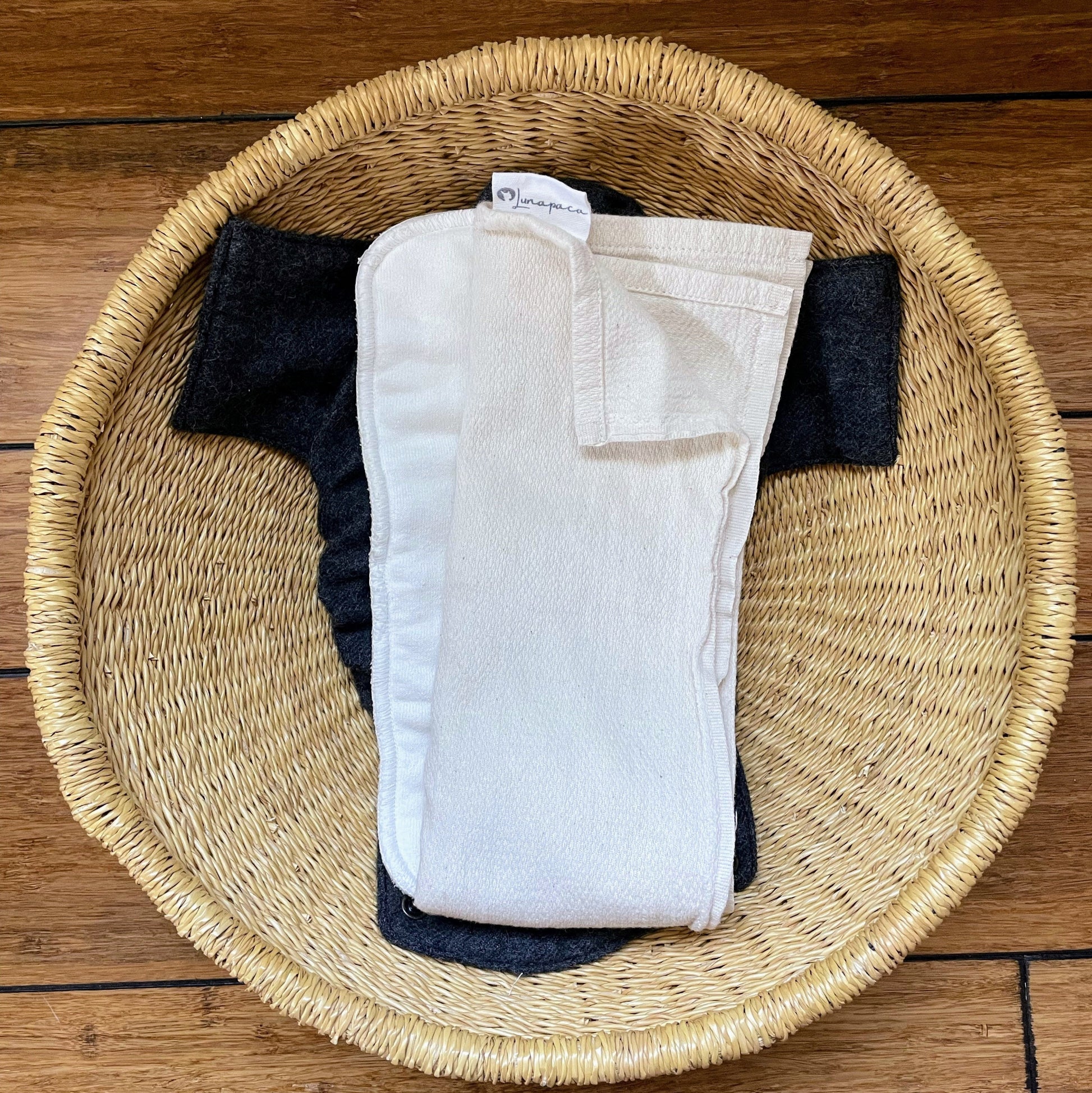 Folded white baby diaper on top of a hemp pad with charcoal alpaca cover in a woven basket on a wooden floor.