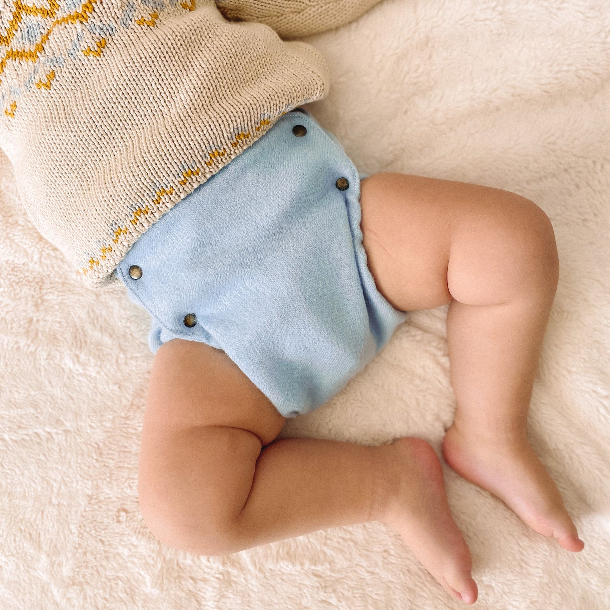 A baby lying on a soft cream-colored rug, wearing a knit sweater and light blue alpaca diaper with brass snaps.
