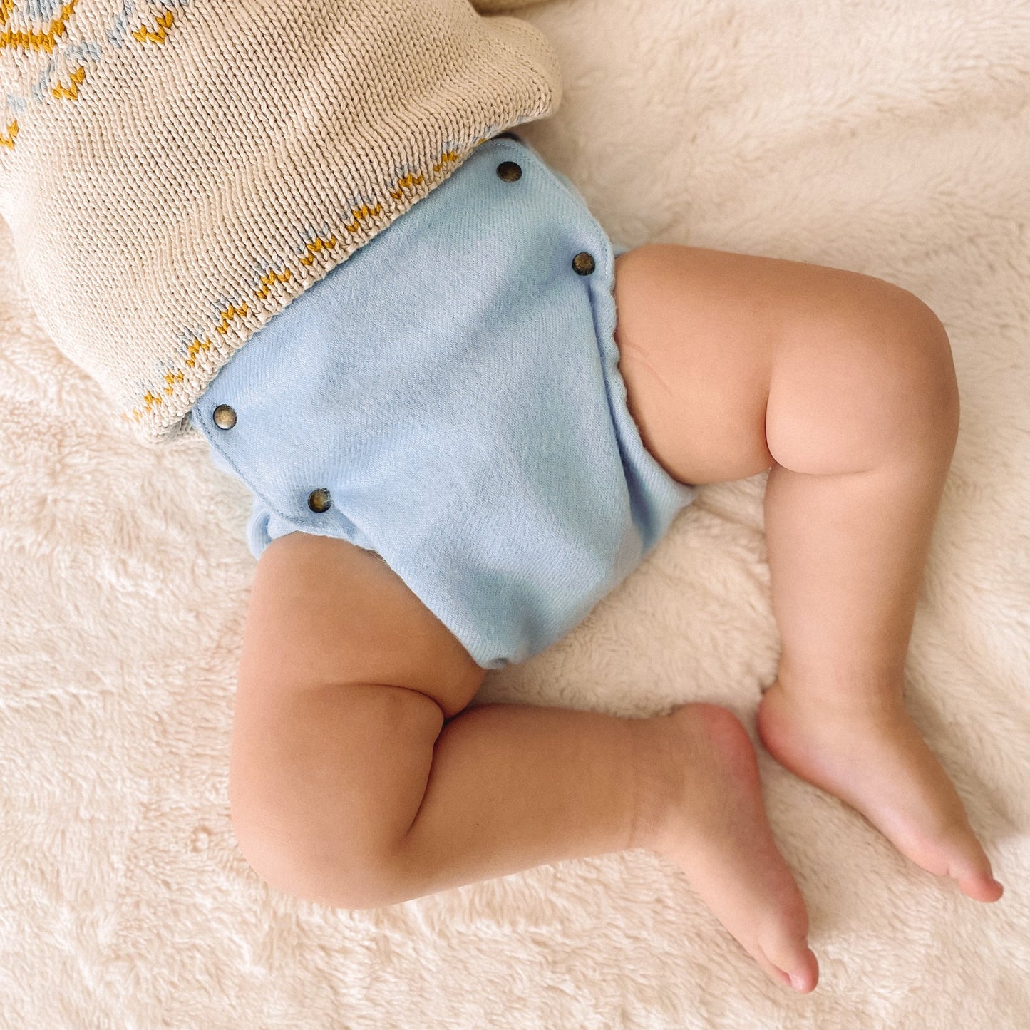 A baby laying on their back on top of a plush white blanket wearing a knitted cream sweater with yellow and blue details and a light blue alpaca diaper cover with brass snaps.