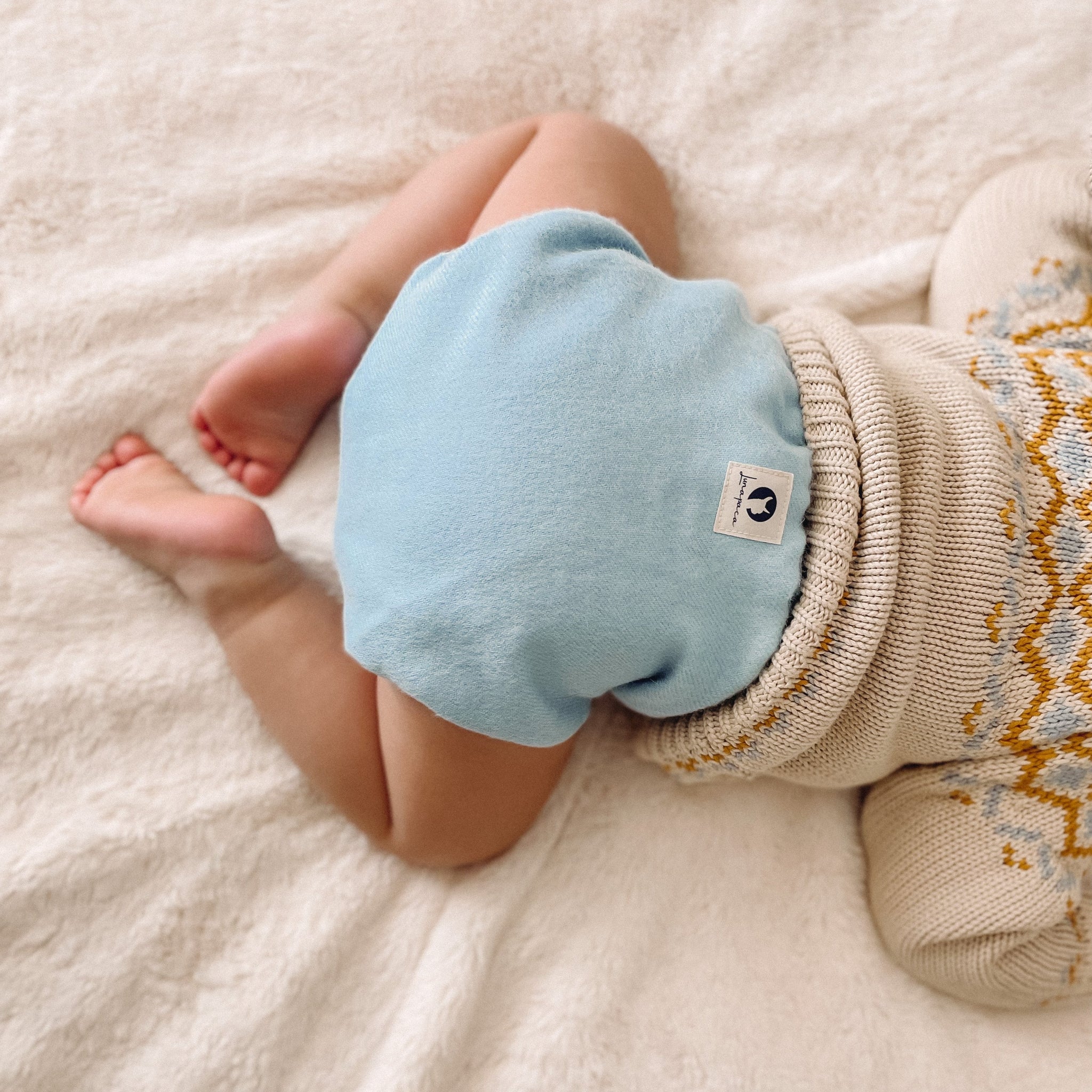 A baby lying on a soft cream-colored blanket, wearing a knit sweater and light blue alpaca diaper.