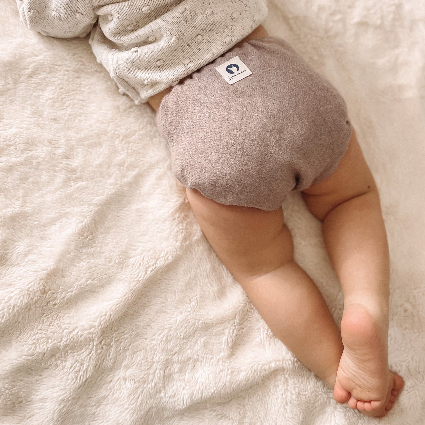 A toddler laying on her stomach on a white blanket wearing a cream colored sweater and a clay colored alpaca diaper cover with a white square logo on the center upper back of the diaper cover.