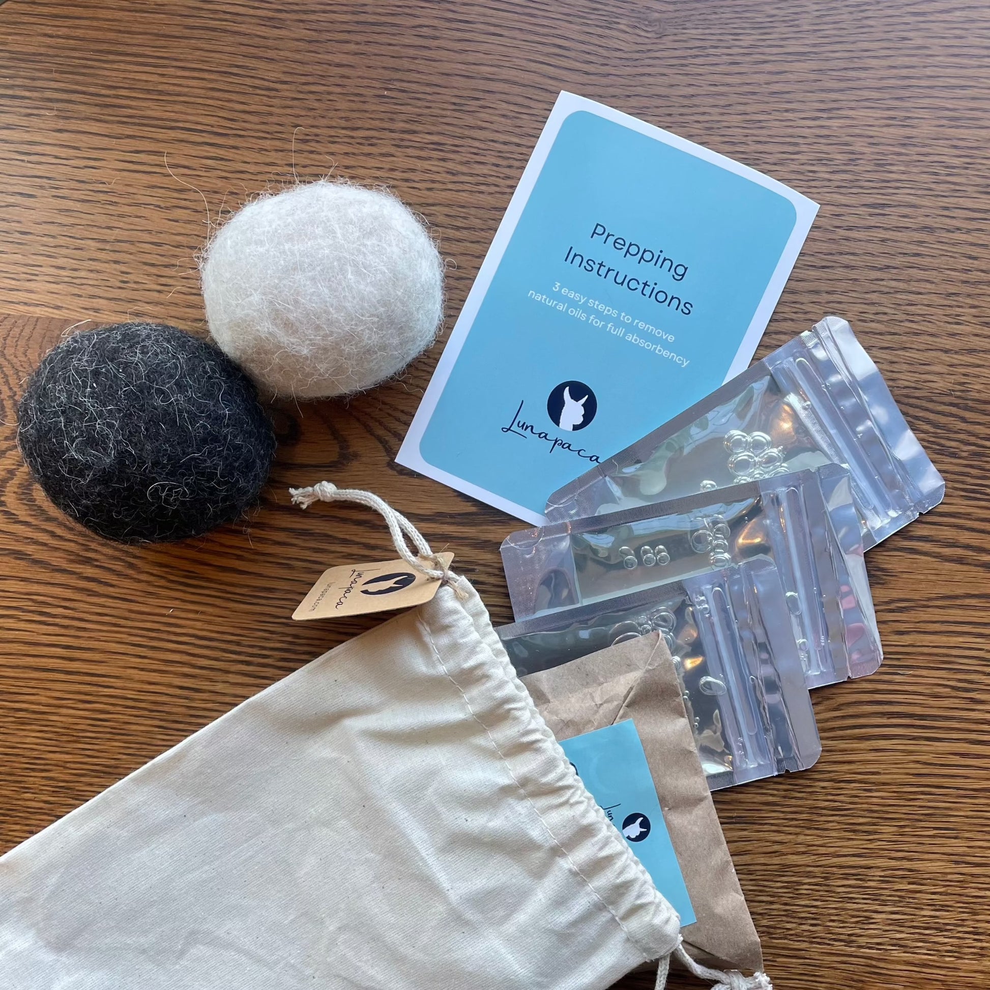 2 alpaca dryer balls next to a card that says "Prepping Instructions" next to 3 packets of liquid detergent, and a sealed paper sack coming out of a cotton drawstring bag on top of a wooden table.