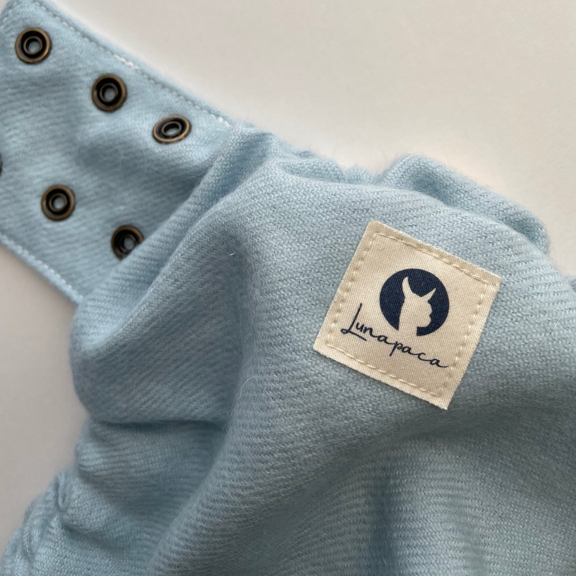 Close-up of a light blue alpaca diaper cover with brass snaps and a sewn-on white Lunapaca brand patch with a white background.