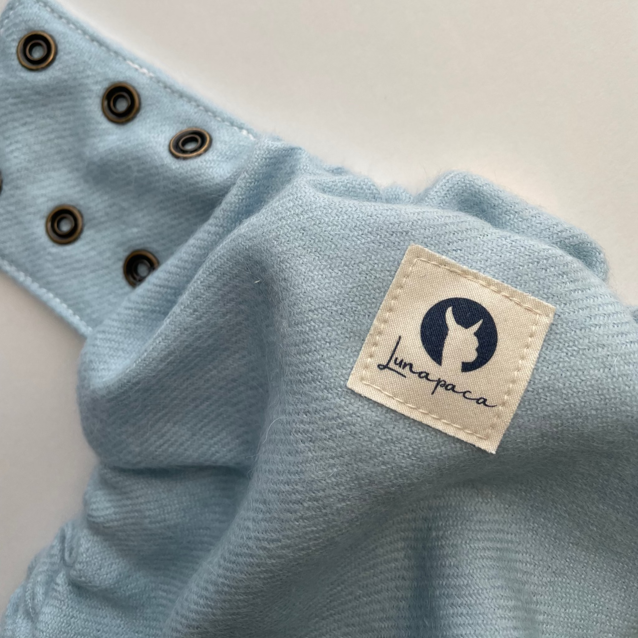 Close-up of a light blue alpaca diaper cover with brass snaps and a sewn-on white Lunapaca brand patch with a white background.