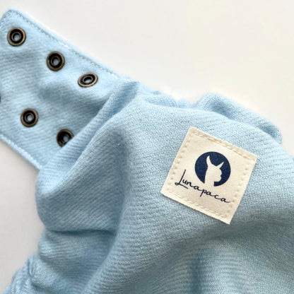 The upper back and side panel of a light blue alpaca diaper cover on white background. A white cotton square logo featuring a navy silhouette of alpaca is in the foreground and brass rings along the side panel are in the foreground.