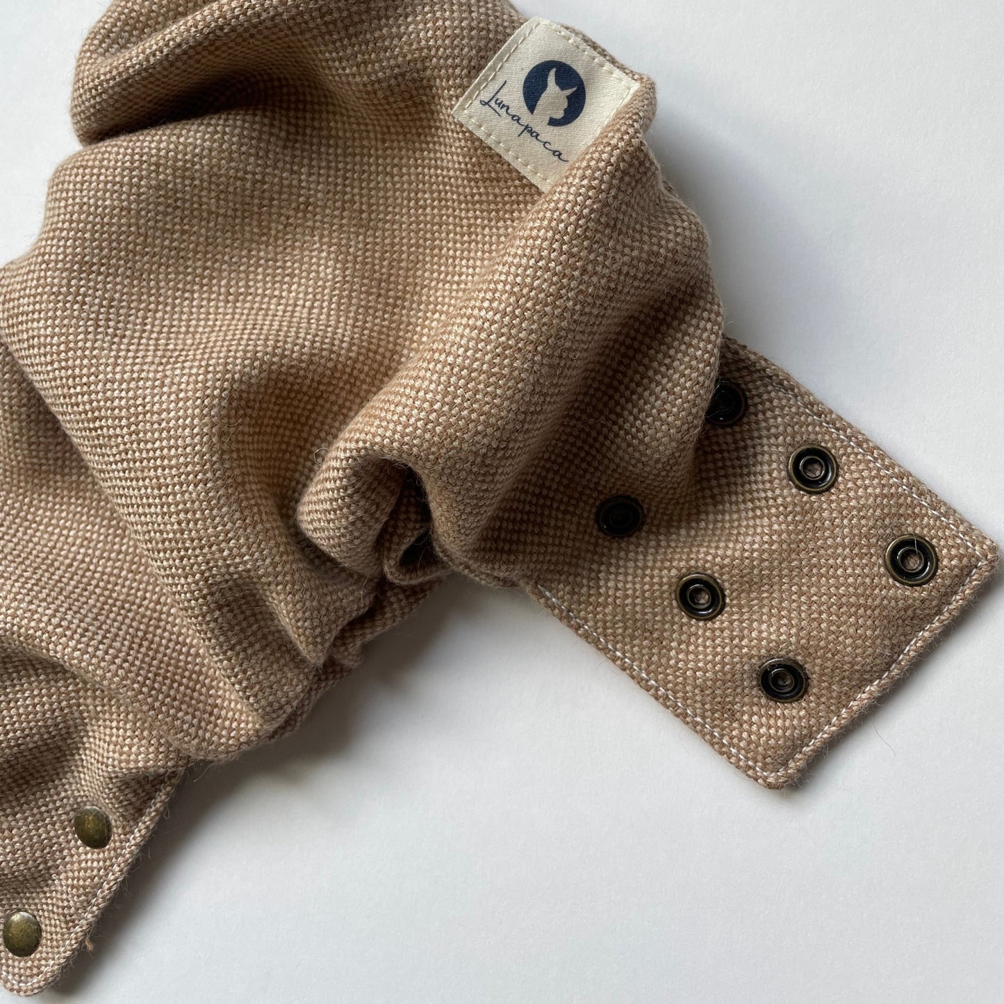 An alpaca diaper cover with a plain weave of light brown and white color lays open on a white background. A white square logo tag featuring an alpaca silhouette and brass snaps along one of the side panels and along the front panel are visible.