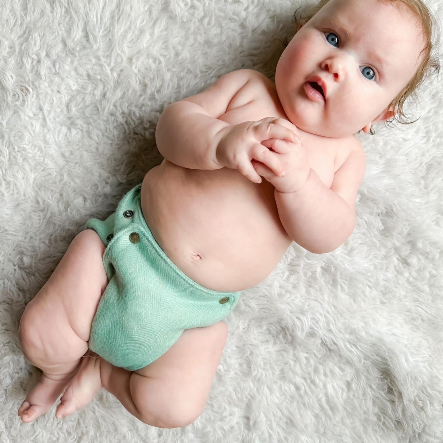 A baby with light colored skin and blue eyes and red hair laying on their back on a white shag carpet wearing a sage green alpaca diaper cover with brass snaps.