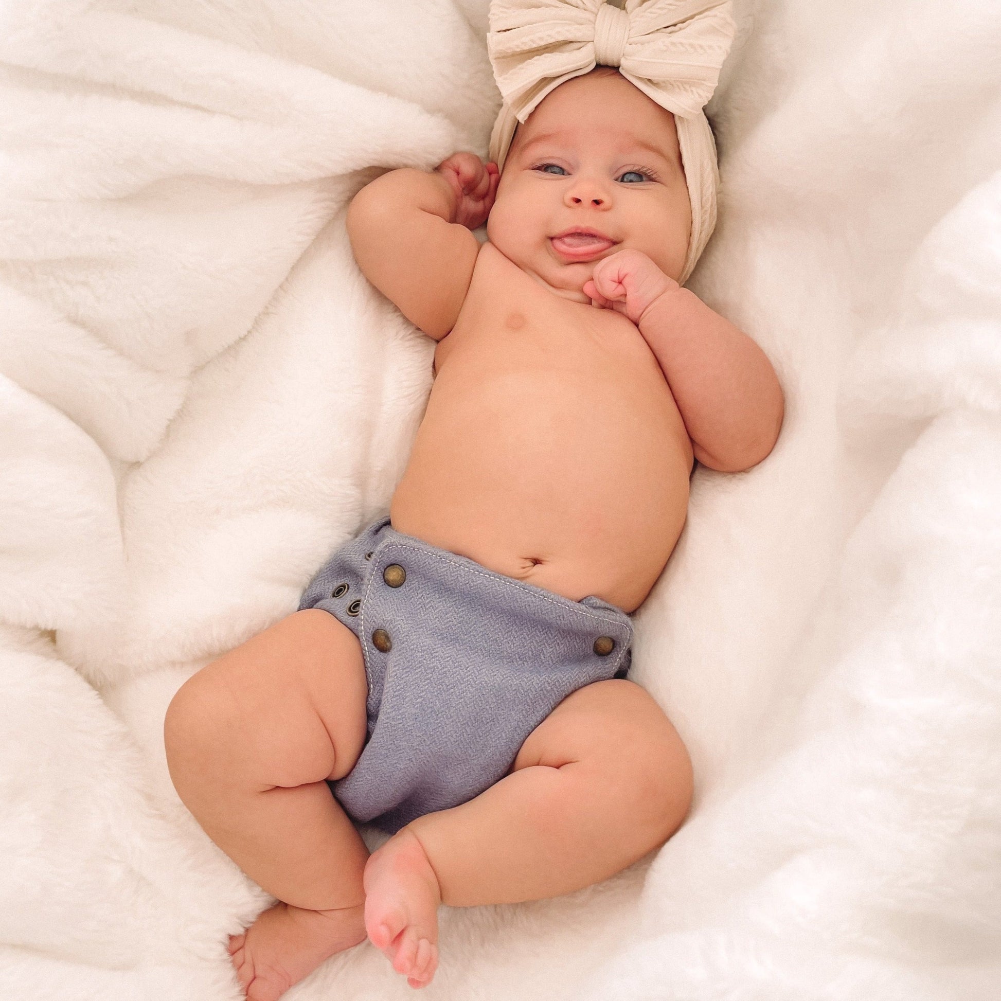 A smiling baby with medium skin tone wearing a cream colored bow around her head plus a dawn-colored alpaca diaper cover with brass snaps. She is laying on top of a plush white blanket.