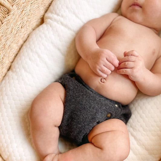 An infant with light skin tone laying in a straw-colored bassinet with a white mattress wearing a charcoal gray alpaca diaper cover.