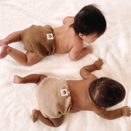 Two babies crawling on top of a plush white blanket. The baby at the top of the photo has medium-dark skin and black hair and is wearing a dark brown alpaca diaper cover. The baby in the bottom of the photo has dark-black skin and black hair and is wearing a light brown diaper cover.