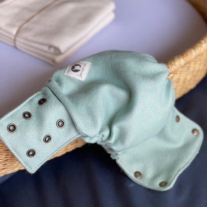 A sage-green alpaca diaper cover with twill weave and brass snaps and square cotton logo tag hangs from the side of a diaper changing basket set on top of a dark blue background. Two folded cotton flat diapers tied with twine is in the background.