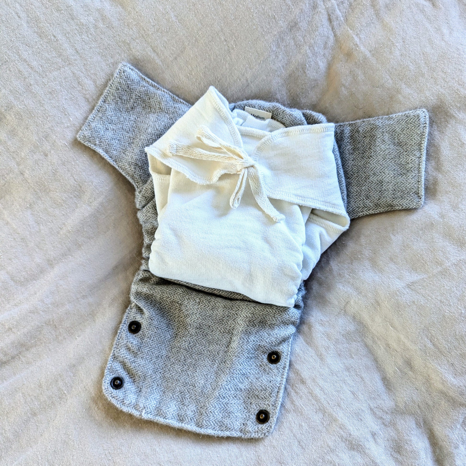 A reusable cloth baby diaper cover in a heather gray woven fabric, shown laid flat on a beige fabric background with a folded cream-colored cotton fitted diaper with a cotton string closure placed on top.