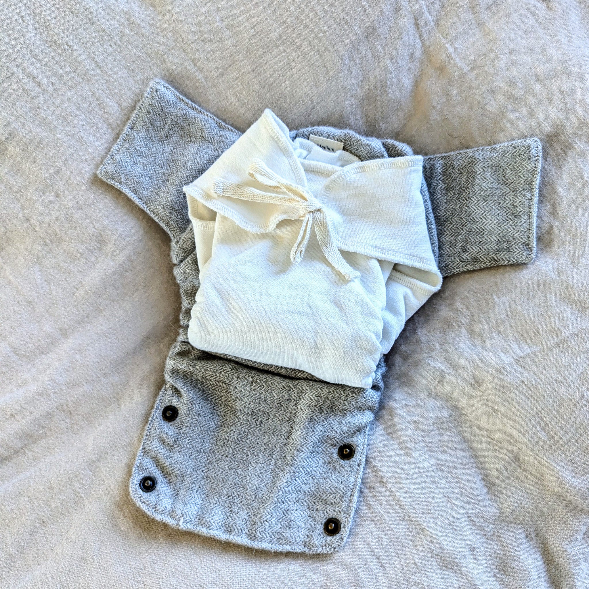 A reusable cloth baby diaper cover in a heather gray woven fabric, shown laid flat on a beige fabric background with a folded cream-colored cotton fitted diaper with a cotton string closure placed on top.