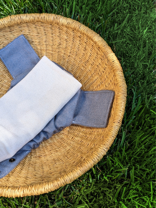 A folded cotton rectangle on top of a unsnapped alpaca diaper cover with a mini herringbone pattern in purple-blue, laying inside a basket on green grass.