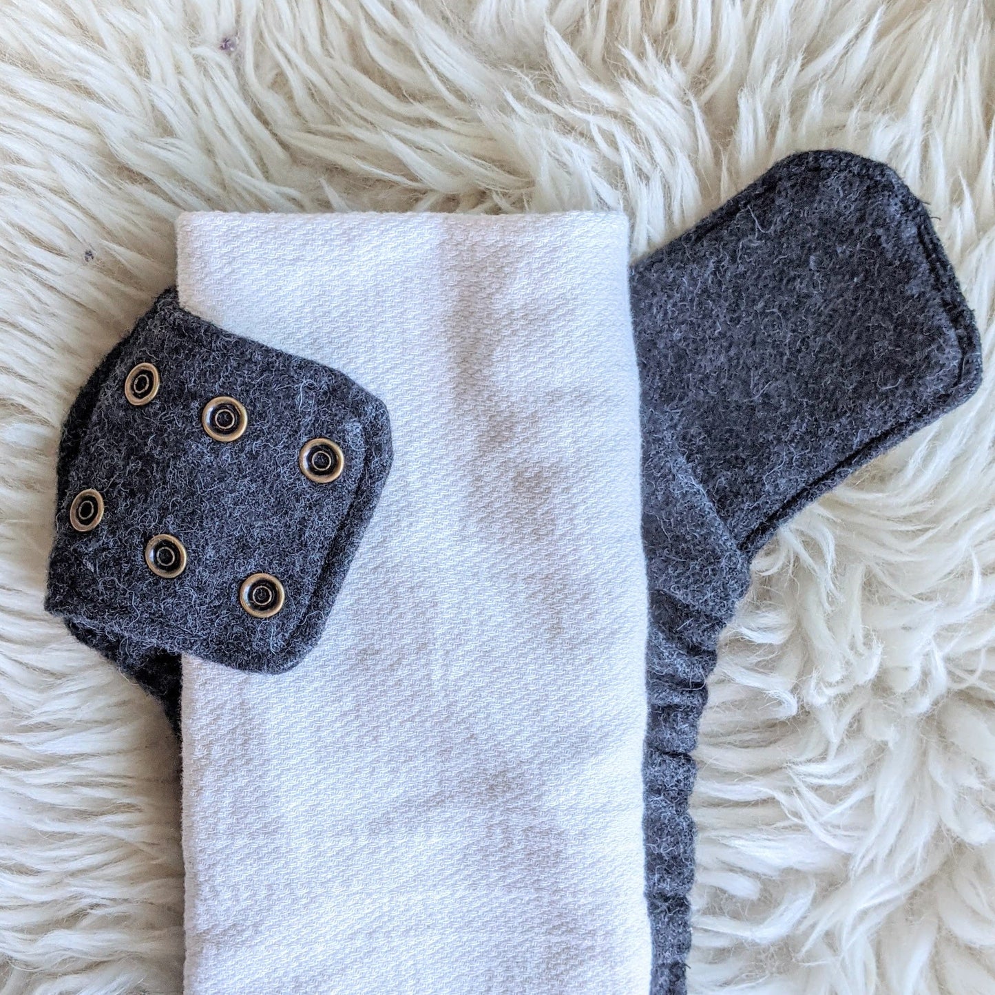 A rectangular "pad-folded" cotton flat diaper on top of a gray alpaca diaper cover with one wing wrapped around the cotton revealing 6 antique brass snaps. The diaper is on top of a fuzzy white sheepskin rug.
